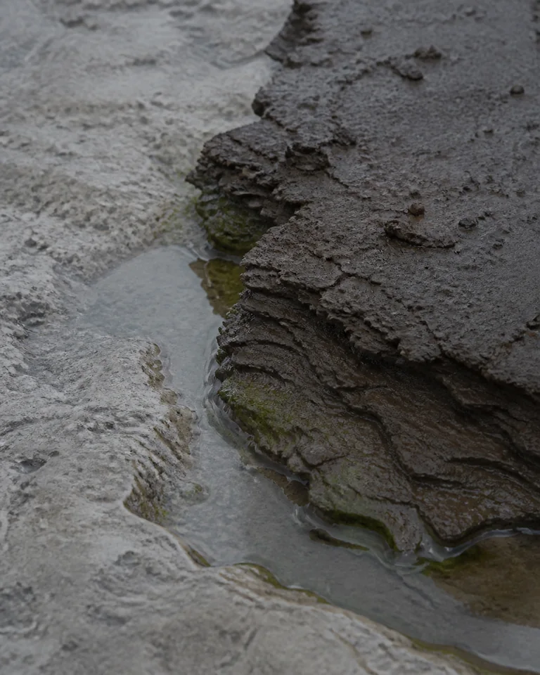 Rain-darkened clay strata meeting cold limestone and a shallow water edge.