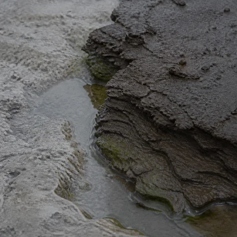 Rain-darkened clay strata meeting cold limestone and a shallow water edge.