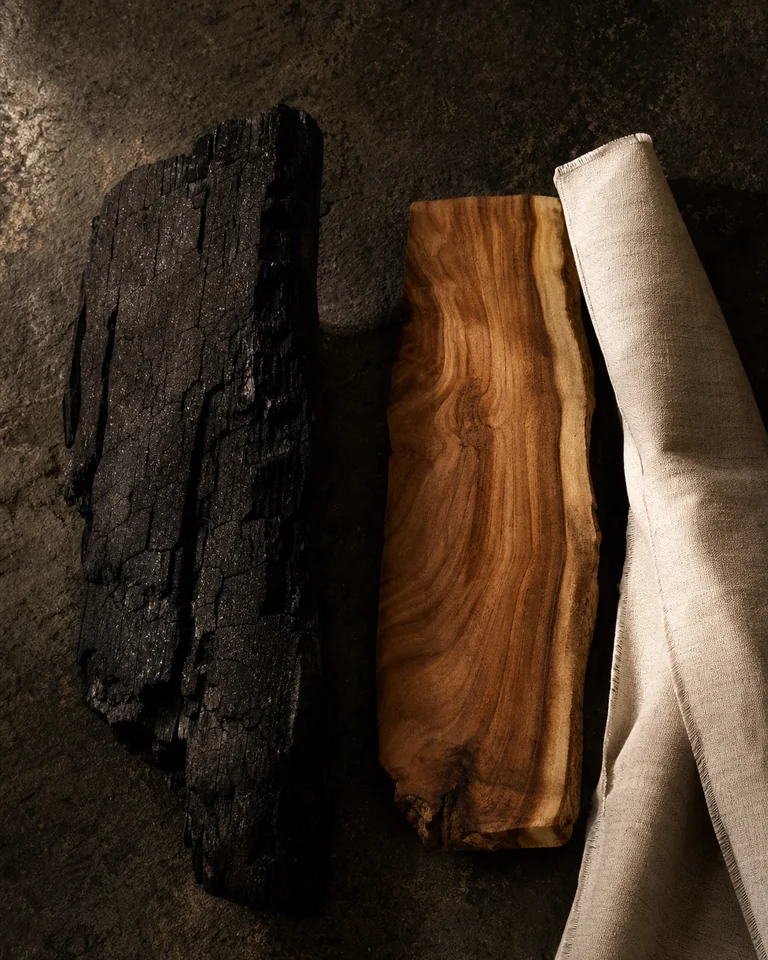 Charred wood beside warm sandalwood grain and bone-coloured linen.