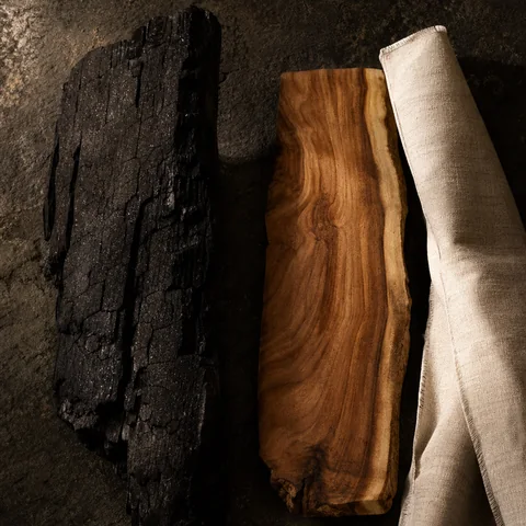 Charred wood beside warm sandalwood grain and bone-coloured linen.